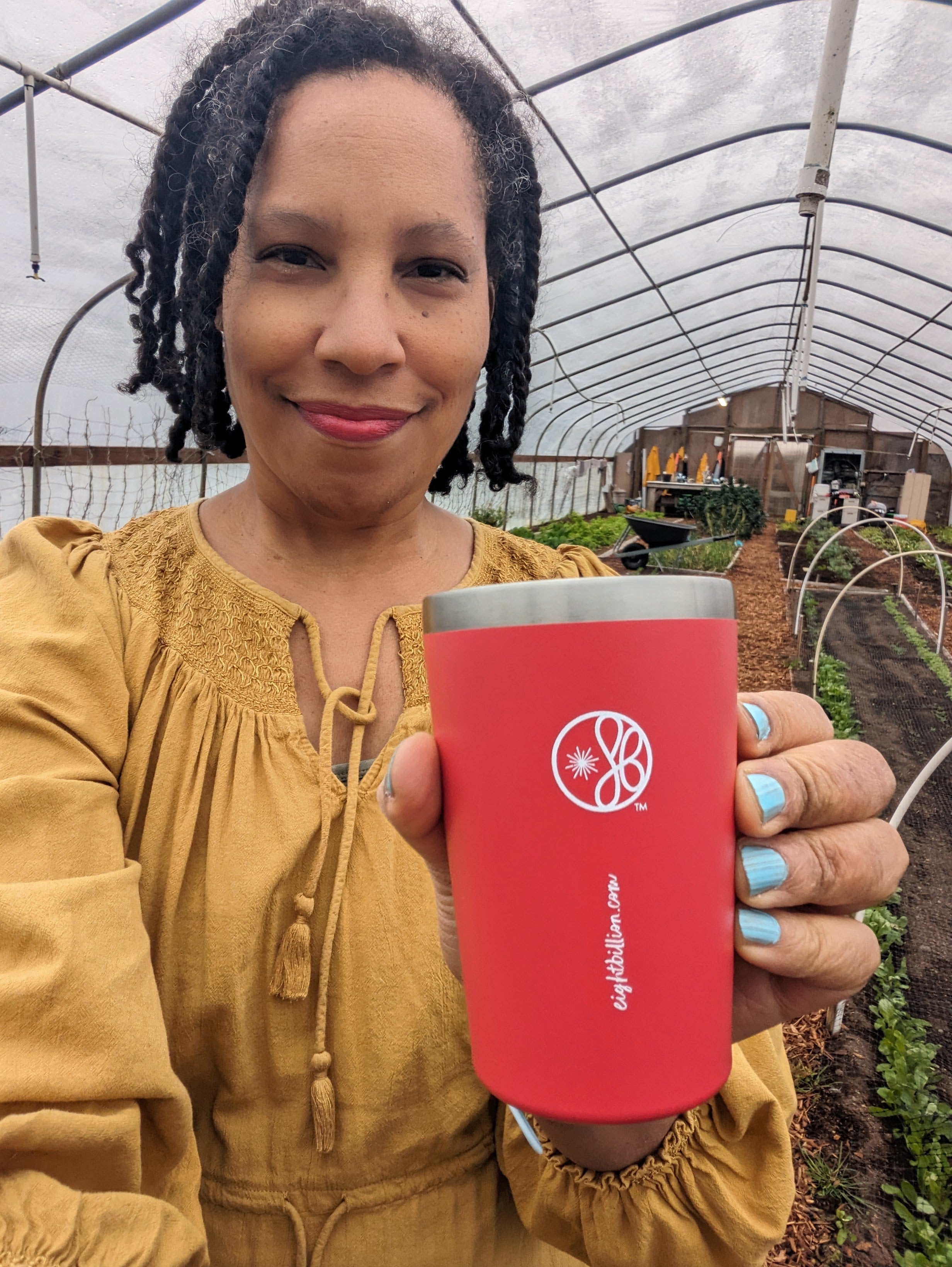 Smiling woman holding an Eight Billion JostArriba red tumbler in a greenhouse. Enjoying a plant-based adaptogenic coffee alternative made with functional mushrooms like lion’s mane and reishi. Natural nootropic drink for focus, mood, and energy—perfect for conscious living and sustainable lifestyles